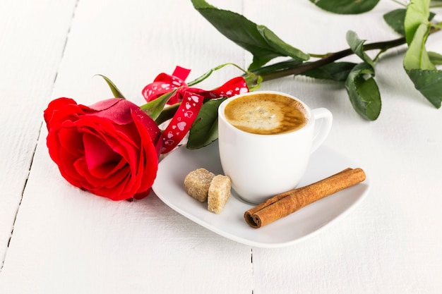 Tazza di caffè, rosa rossa, zucchero e cannella su un fondo di legno bianco