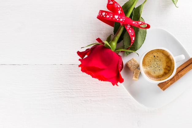 Tazza di caffè, rosa rossa, zucchero e cannella su un fondo di legno bianco