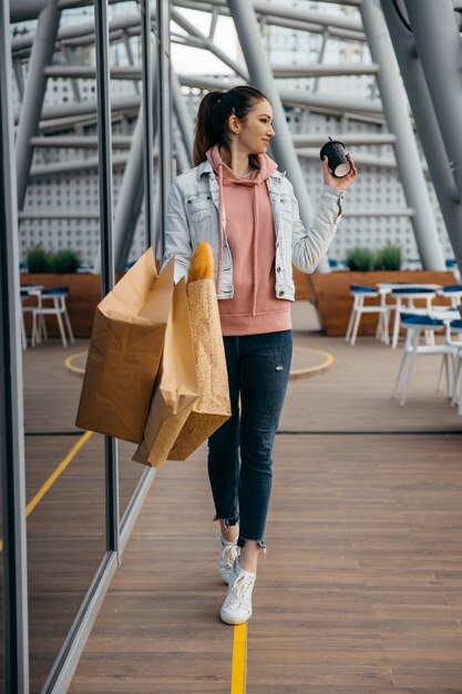 Tazza di caffè sorridente della tenuta della giovane donna, camminante con il sacco di carta di compera della drogheria con le baguette lunghe del pane bianco