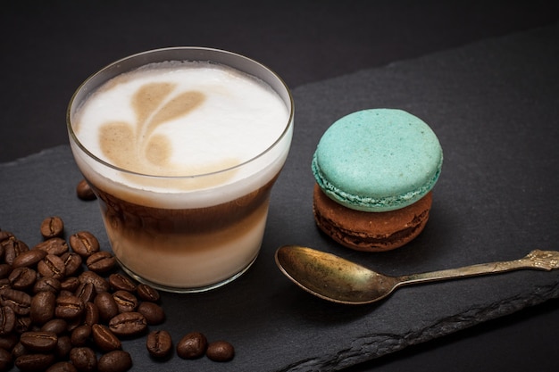 Tazza di cappuccino, chicchi di caffè, cucchiaio e amaretti su sfondo nero. Vista dall'alto.