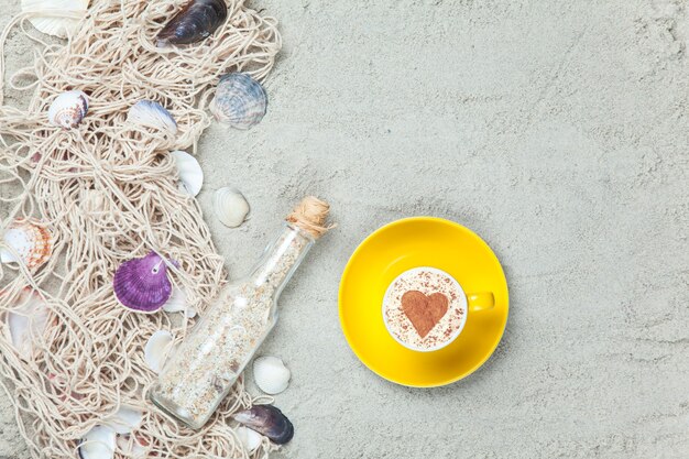 Tazza di cappuccino con simbolo a forma di cuore, bottiglia e rete con conchiglie sulla sabbia