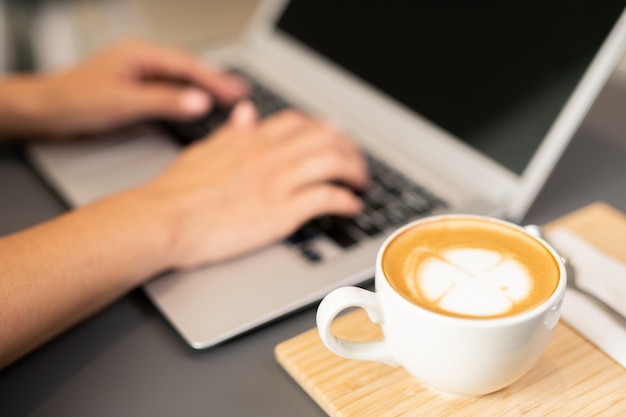 Tazza di cappuccino sul vassoio in legno accanto alle mani della studentessa toccando la tastiera del computer portatile durante la rete nella caffetteria