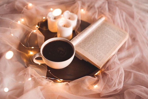 Tazza di cioccolata calda con candele e libro aperto che resta sul vassoio di legno nel primo piano del letto. Buon giorno. Colazione. Fare una pausa caffè.