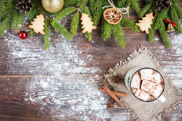 Tazza di cioccolata calda con marshmallow, spezie, biscotti allo zenzero di Natale, rami di abete e coni su fondo di legno scuro. Copia spazio per il testo. Carta da parati natalizia, carta.