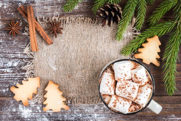 Tazza di cioccolata calda con marshmallow, spezie, biscotti allo zenzero di Natale, rami di abete e coni su fondo di legno scuro. Copia spazio per il testo. Carta da parati natalizia, carta.