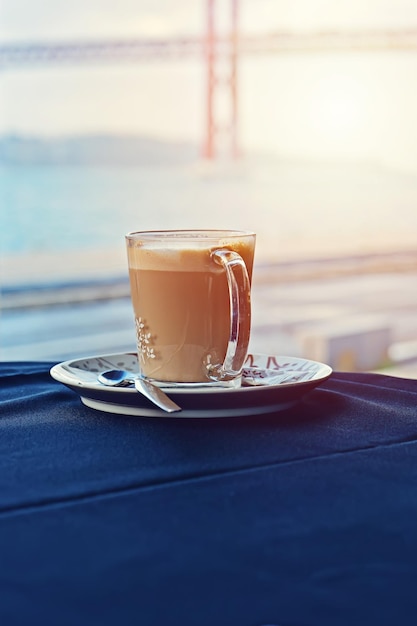 Tazza di latte caldo sul tavolo con tovaglia blu contro la finestra con il ponte di Lisbona