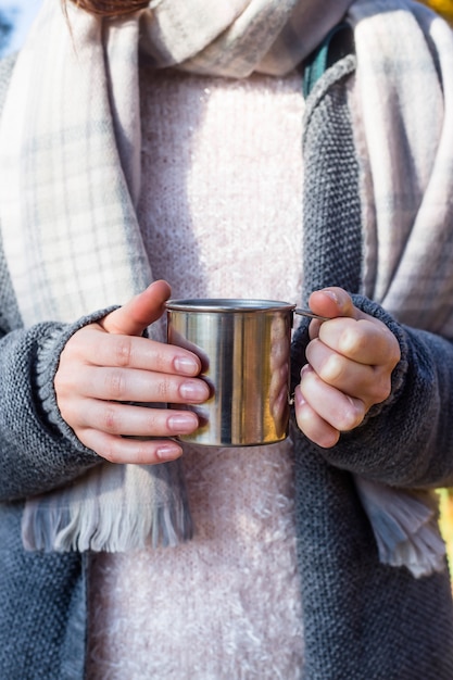 Tazza di metallo con una bevanda calda in mani femminili. Vestiti caldi autunnali lavorati a maglia