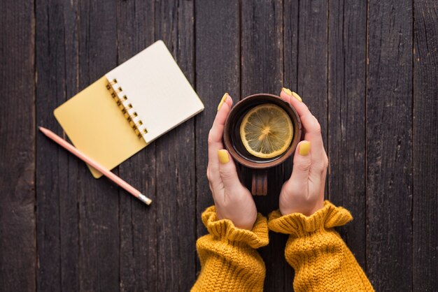 Tazza di tè al limone in una mano femminile, matita e blocco note. Fondo in legno. Vista dall'alto