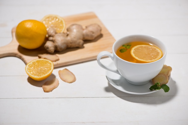 Tazza di tè allo zenzero con limone e miele su uno sfondo di legno bianco.