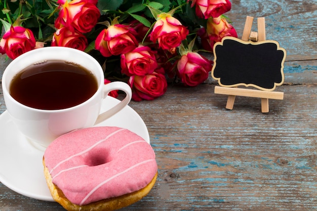 Tazza di tè con ciambella, rose fresche e lavagna con spazio per il testo su uno sfondo di legno. Immagine perfetta per la festa della mamma.