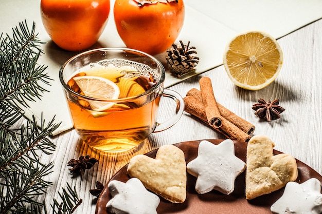 Tazza di tè con fetta di limone, biscotti di panpepato a forma di stella e cuore, rami di abete naturale con cono e cannella, anice stellato su tavole di legno. Effetto tonificante del colore.