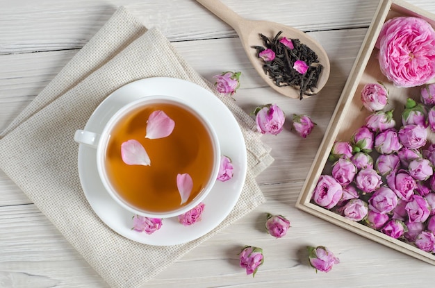 Tazza di tè con pani e boccioli di rosa e cucchiaio di tè sulla tavola di legno bianca.