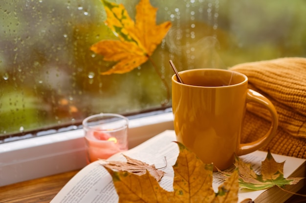 Tazza di tè, libro, foglie autunnali e candela sul davanzale della finestra a casa