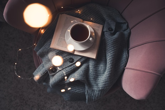 Tazza di tè nero fresco che rimane su un libro di carta aperto con un panno di tessuto a maglia su una comoda sedia d'accento da vicino. Buon giorno. Ora di colazione.