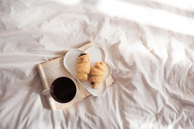 Tazza di tè nero fresco con croissant sul piatto che resta sul libro di carta aperto a letto in primo piano della luce del sole. Vista dall'alto Buongiorno. Ora di colazione.