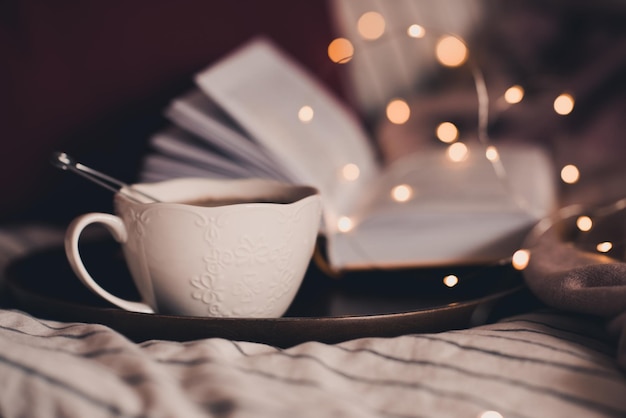 Tazza di tè nero su vassoio di legno con libro aperto e luci incandescenti al primo piano di sfondo. Buon giorno. Ora di colazione. Pausa caffè.