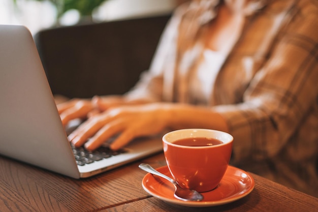 Tazza di tè rossa sullo sfondo della donna in camicia a quadri che lavora con il computer portatile al caffè