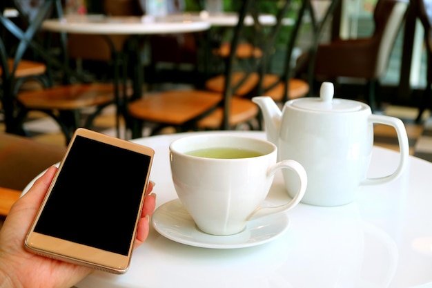 Tazza di tè verde caldo e teiera sul tavolo del caffè con la mano femminile che tiene un telefono cellulare