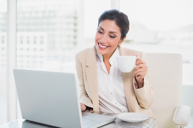 Tazza di tenuta felice della donna di affari mentre scrivendo sul computer portatile