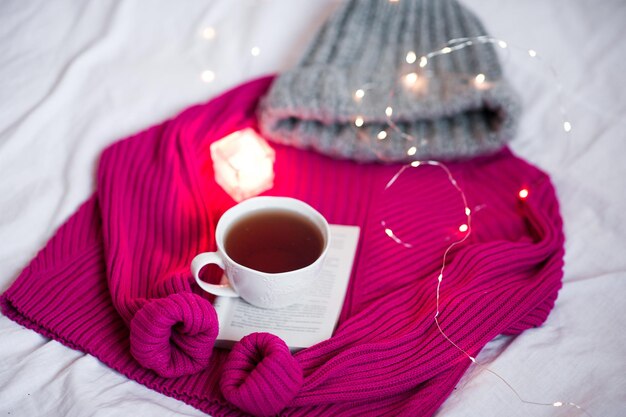 Tazza di tes nero su maglione lavorato a maglia con candele e cappello intrecciato in primo piano a letto. Stagione invernale.