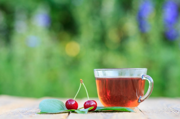 Tazza di vetro di tè e frutti di ciliegia matura rossa con peduncoli su vecchie tavole di legno in sfondo naturale sfocato