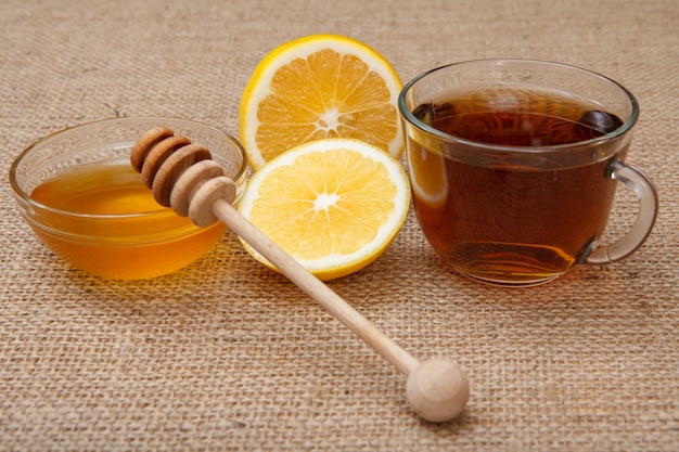 Tazza di vetro di tè, pezzi di limone e miele in una ciotola di vetro con tavola di legno cucchiaio con sacco.