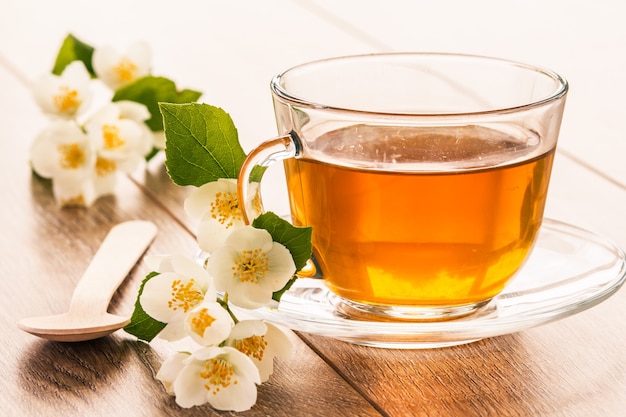 Tazza di vetro di tè verde con fiori di gelsomino bianco e cucchiaio su assi di legno