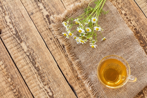 Tazza di vetro di tè verde e fiori di camomilla bianchi freschi su tela di sacco e vecchio fondo di legno. Vista dall'alto.