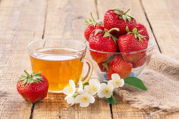 Tazza di vetro di tè verde e fragole mature rosse in una ciotola di vetro con fiori di gelsomino su tela di sacco e tavole di legno