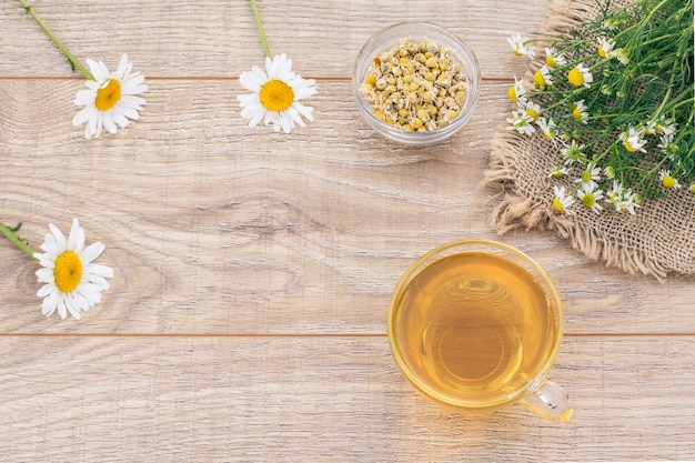 Tazza di vetro di tè verde, fiori freschi di camomilla e una piccola ciotola di vetro con fiori secchi di matricaria chamomilla sullo sfondo di legno. Vista dall'alto.