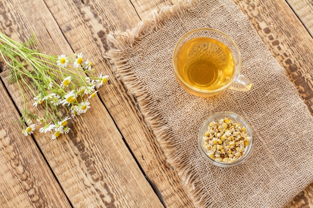 Tazza di vetro di tè verde, piccola ciotola di vetro con fiori secchi di matricaria chamomilla su tela di sacco e fiori di camomilla bianchi freschi su fondo di legno. Vista dall'alto.