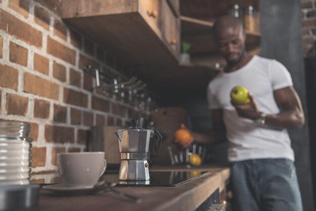 Tazza e caffettiera sul tavolo della cucina e uomo afroamericano in piedi dietro con frutta