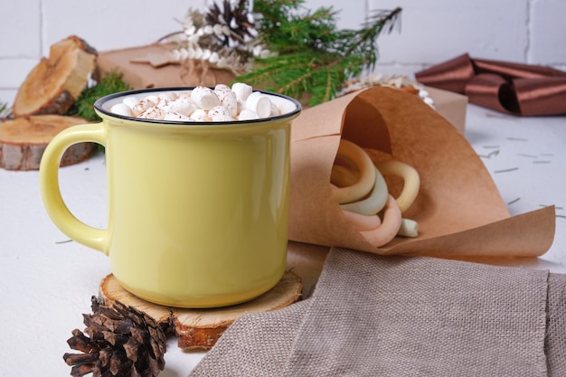 Tazza gialla con cacao decorata con marshmallow su un supporto in legno tagliato a sega, una borsa con marshmallow, un ramo di abete e regali di Natale sullo sfondo