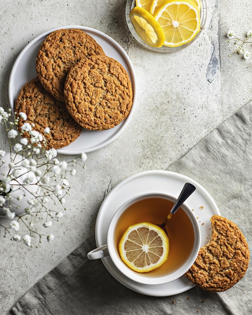 Tazza in porcellana bianca con tè al limone e biscotti di farina d'avena su sfondo strutturato con tovaglia e fiori bianchi. Vista dall'alto