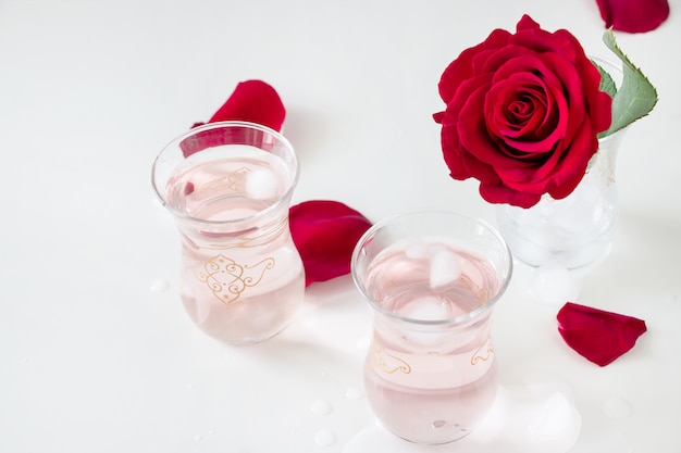 tazza Rinfrescante limonata di acqua di rose con cubetti di ghiaccio e fiore di rosa in vetro e petali su bianco