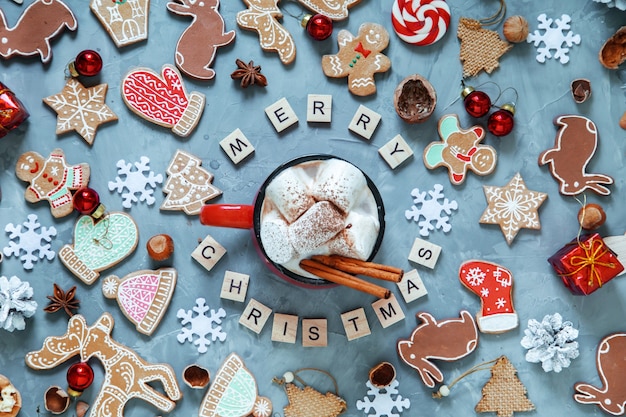 Tazza rossa con cacao e marshmallow, pan di zenzero natalizio e la scritta "Merry Christmas" in lettere di legno.