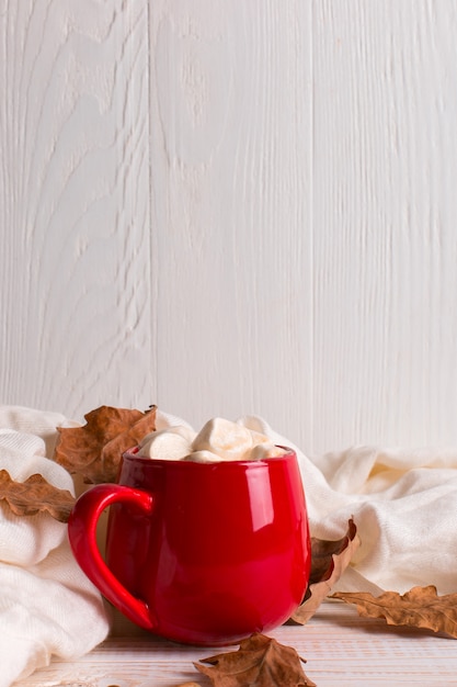 Tazza rossa con cacao e marshmallows, su uno sfondo di una sciarpa e foglie secche. Mood autunnale, una bevanda calda.