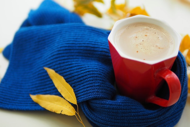 Tazza rossa con caffè sul tavolo in una sciarpa lavorata a maglia blu e foglie autunnali gialle. ciao concetto d'autunno. malattie stagionali