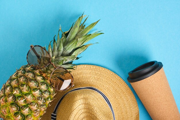 Tazza termica, ananas in occhiali da sole e cappello di paglia su sfondo blu
