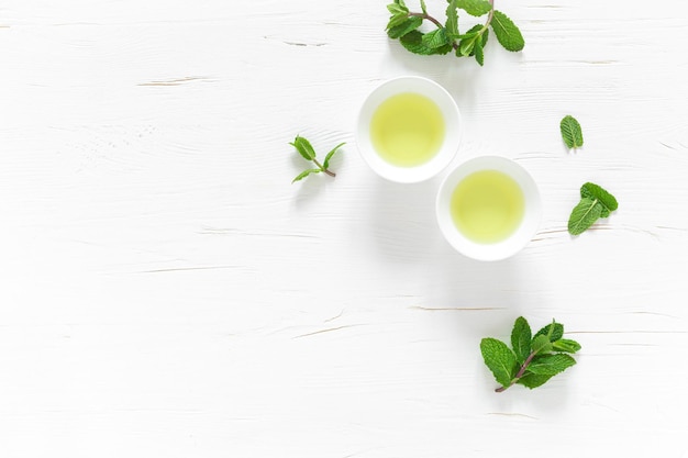 Tè alla menta verde con foglie fresche in tazze e teiera sopra la tavola di legno bianca bevanda riscaldante sana bevanda antiossidante vista dall'alto