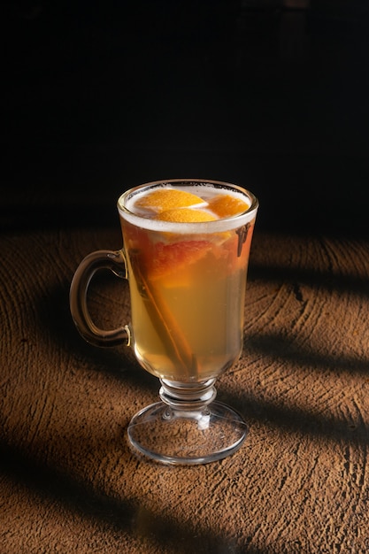 Tè caldo alla frutta con fette d'arancia, chiodi di garofano e bastoncini di cannella in un bicchiere di vetro trasparente con manico.