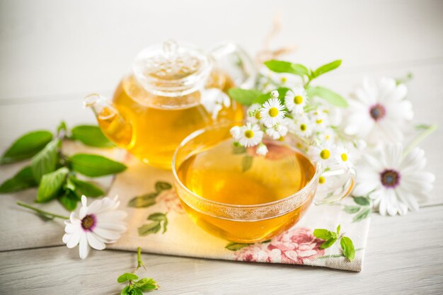 Tè di fiori di camomilla in una tazza di vetro e teiera su un tavolo di legno chiaro