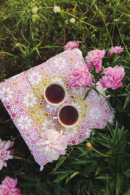 Tè in stile country nel giardino estivo del villaggio Due tazze di tè nero caldo su tovaglia di pizzo vintage lavorata all'uncinetto e fiori di peonia in fiore alla luce del sole