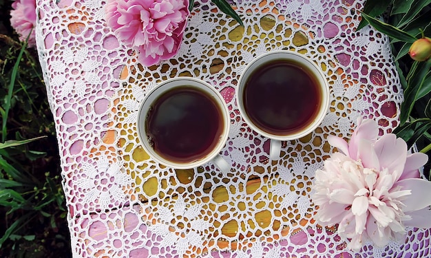 Tè in stile country nel giardino estivo Due tazze di tè nero su tovaglia di pizzo vintage fatta a mano all'uncinetto e fiori di peonia in fiore alla luce del sole