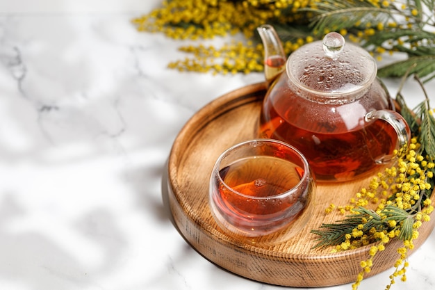 Tè in tazza di vetro e teiera di vetro su vassoio di legno con mimosa Luce solare Spazio di copia