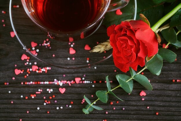 Tè in una tazza trasparente, caramelle di colore e una rosa rossa sul buio