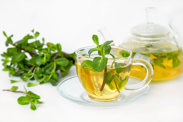 Tè verde alla menta in una tazza e una teiera in vetro trasparente su un tavolo bianco.