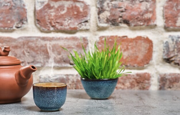 Tè verde caldo in una ciotola blu, fuoco selettivo, vapore sale sopra la tazza, accanto a una teiera di argilla. Tavolo in pietra grigia, muro vintage in mattoni. Primo piano, cerimonia del tè, minimalismo, posto per il testo