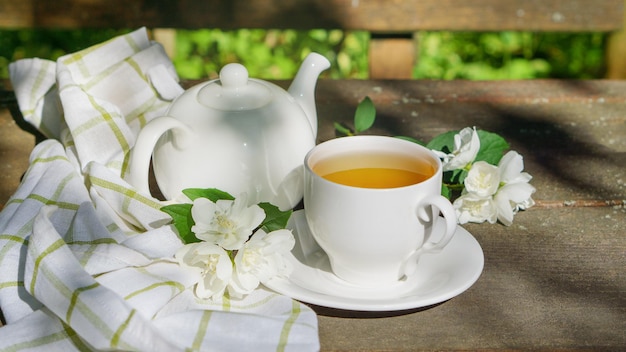 Tè verde caldo in una teiera e una tazza con un ramo di fiori di gelsomino sbocciano e asciugamano bianco su fondo di legno marrone rustico ruvido. Concetto sano di vita lenta della natura.