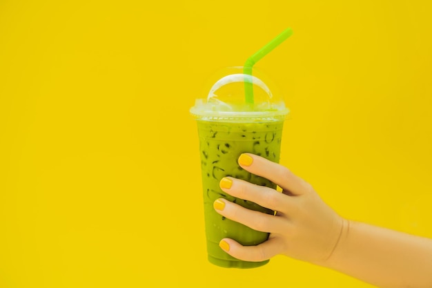 Tè verde latte con ghiaccio in tazza di plastica e cannuccia in mano femminile con manicure gialla su sfondo giallo Tè freddo Matcha Latte fatto in casa con latte da asporto